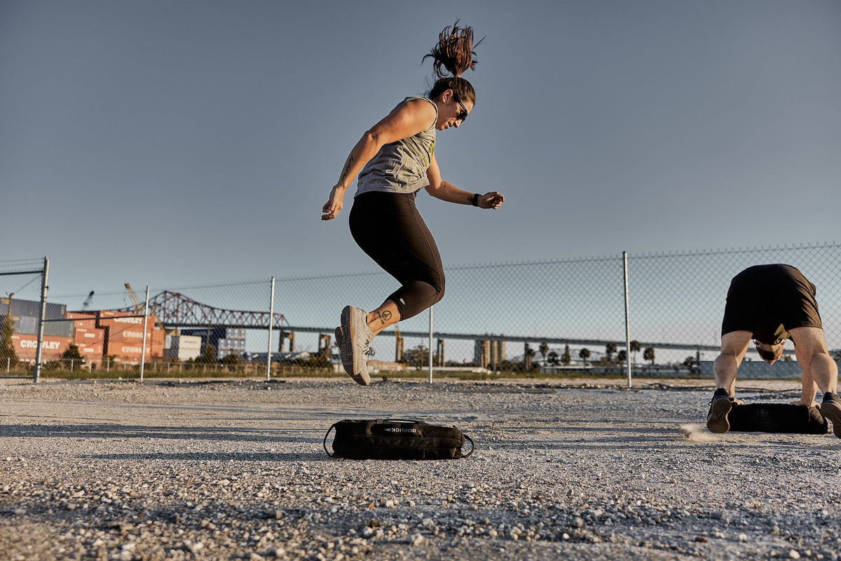Woman jumping over GORUCK sandbag outdoors during rucking workout with shipping containers in background.
