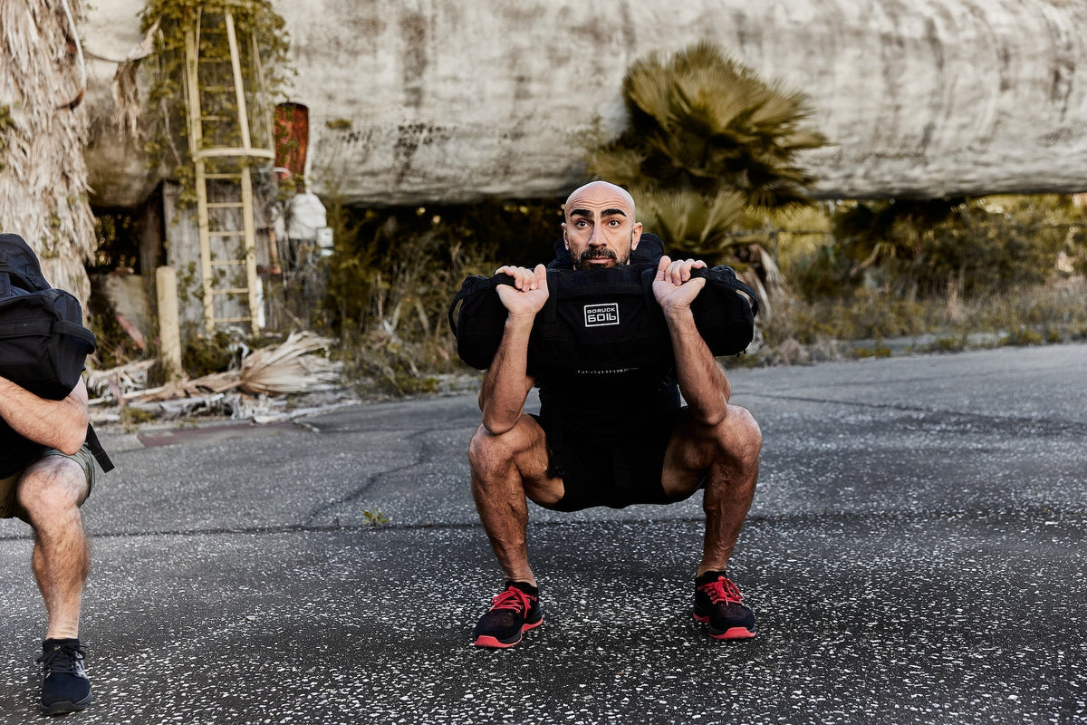 Man performing outdoor squat with GORUCK rucking sandbag, wearing athletic gear