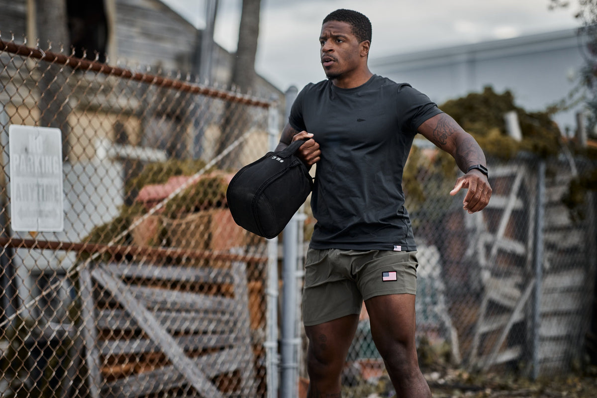 Man training outdoors with GORUCK sandbag, wearing black shirt and olive shorts with US flag patch