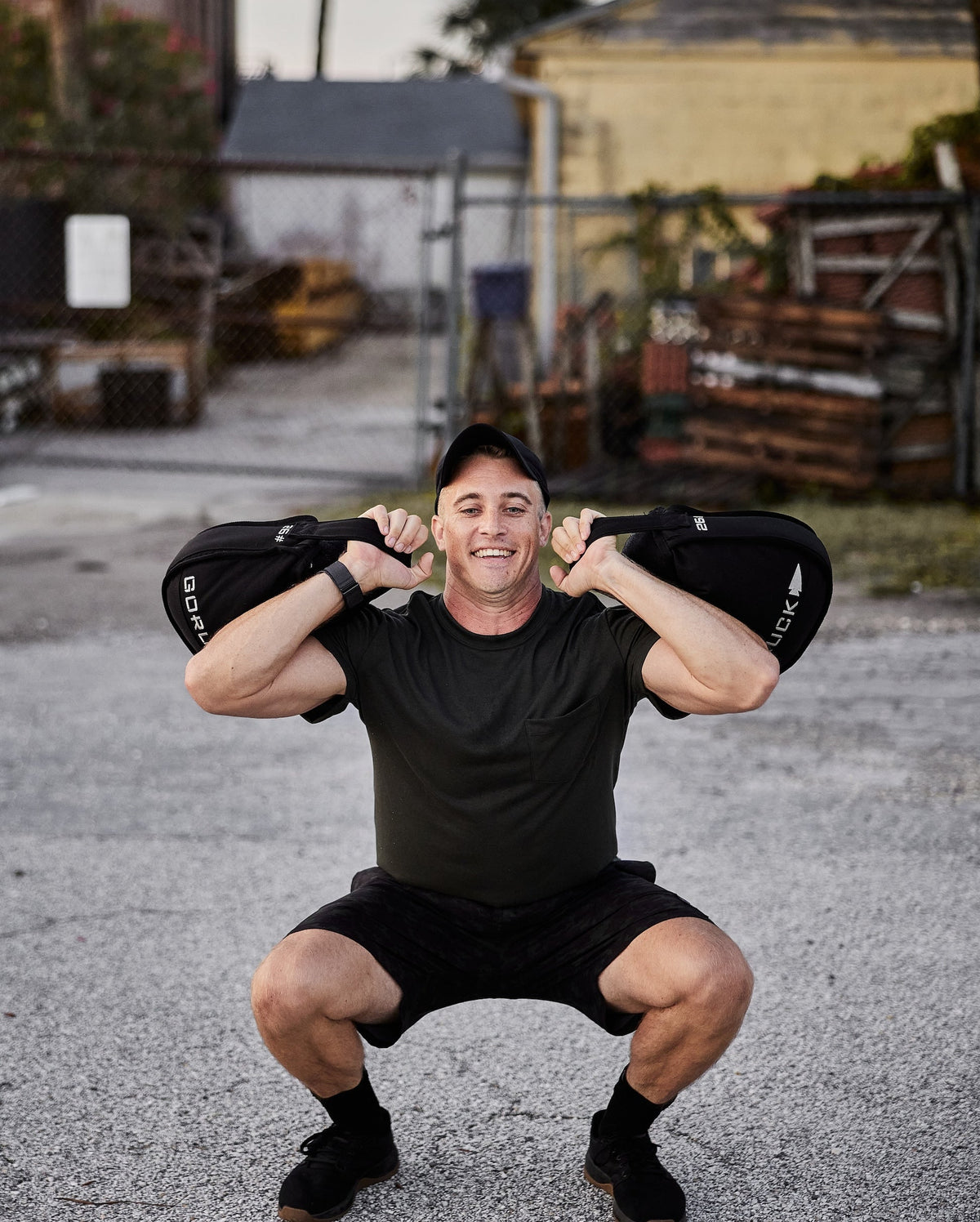 Man doing outdoor squat with GORUCK sandbag, wearing black athletic gear, urban background