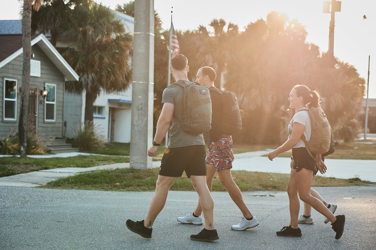 Three people rucking with GORUCK backpacks outdoors in sunlight, walking near houses