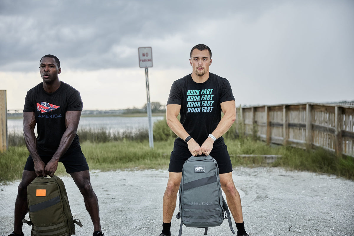 Two men rucking outdoors with GORUCK fitness backpacks, standing on a gravel path by water.