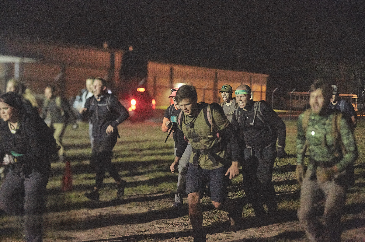 Group of people wearing backpacks rucking at night on grass, GORUCK endurance event