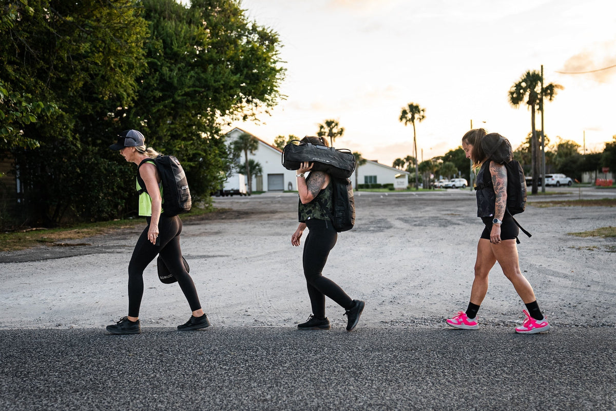 Three women rucking with GORUCK gear outdoors at sunrise, carrying weighted backpacks and sandbags.
