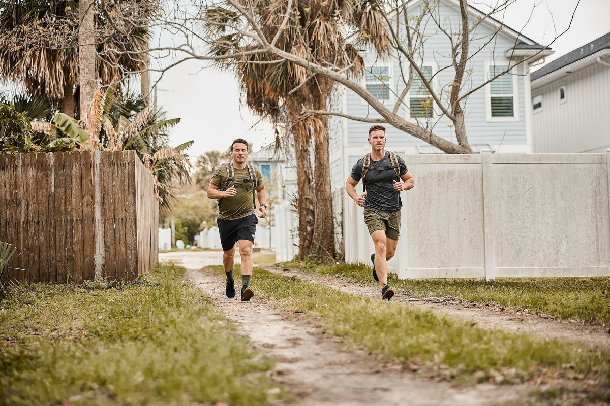 Two men rucking outdoors with GORUCK backpacks on a dirt path by palm trees and houses