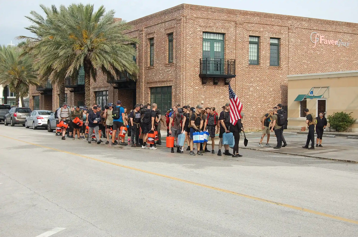 Group of people rucking with GORUCK gear, carrying buckets and American flag on city street