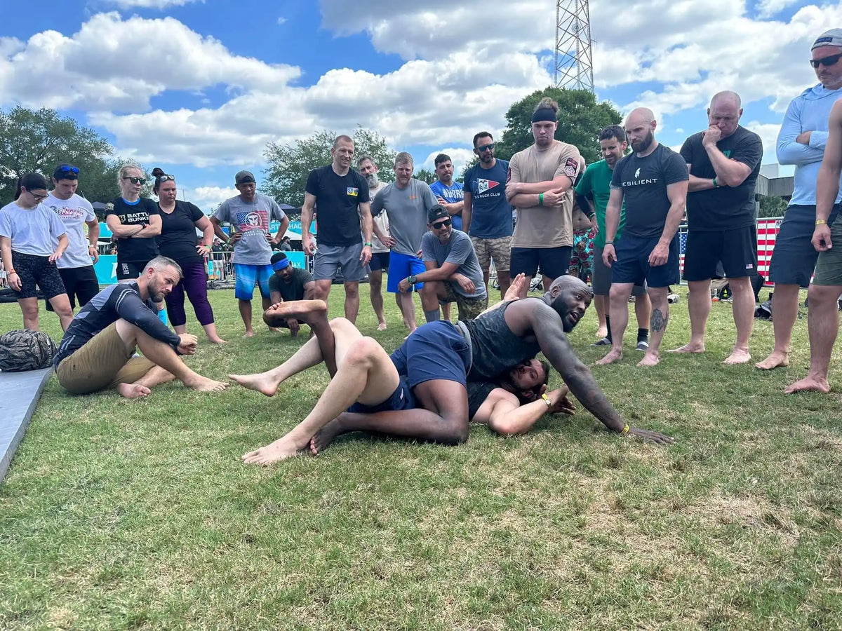 Outdoor GORUCK event with people demonstrating grappling techniques on grass, group watching.