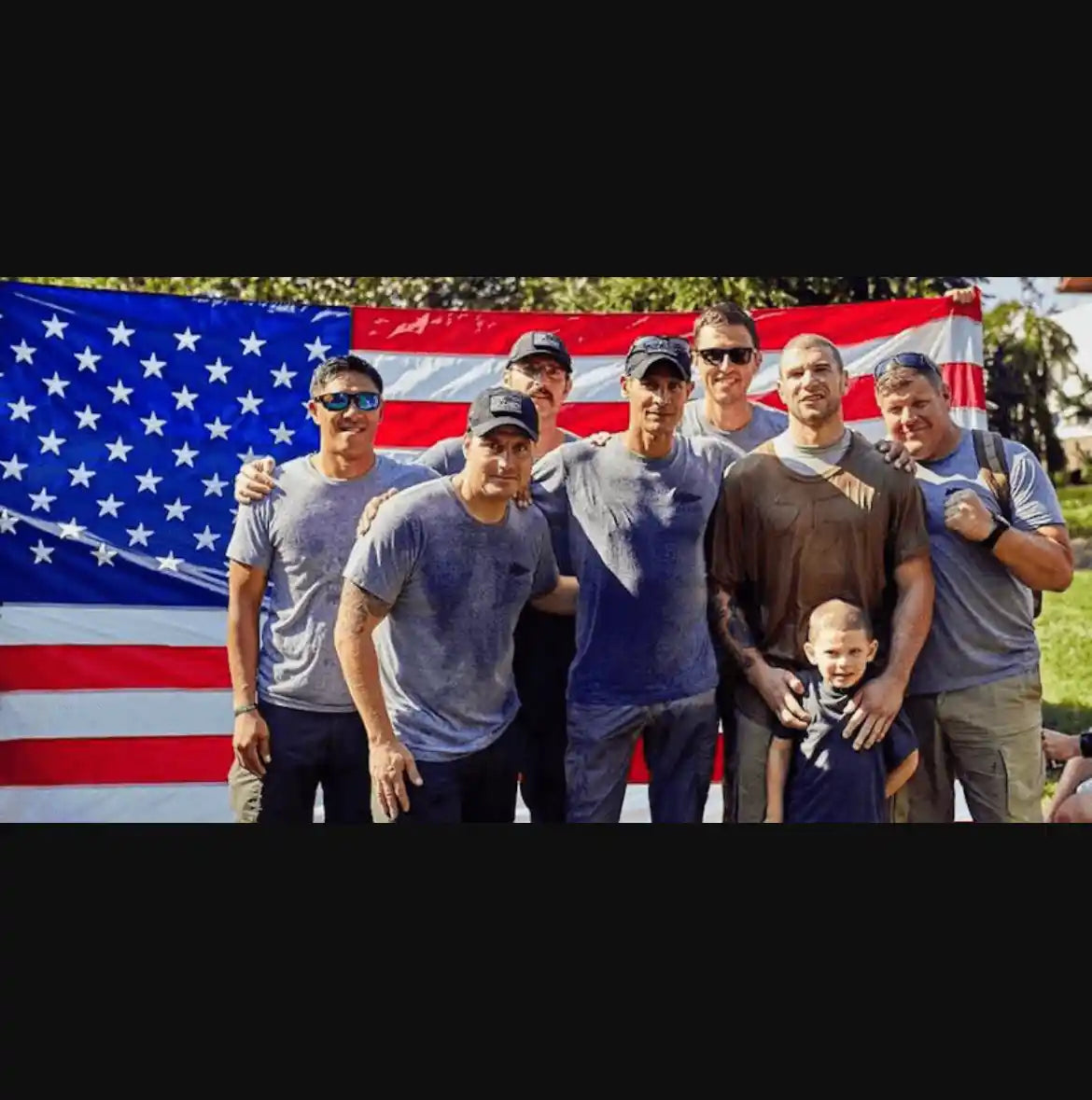 Group of men in GORUCK gear with a child, posing in front of large American flag outdoors