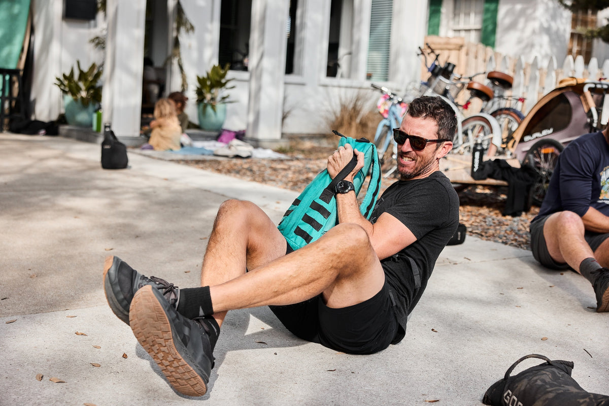 Man outdoors doing sit-ups with GORUCK rucksack, wearing black athletic gear and sunglasses