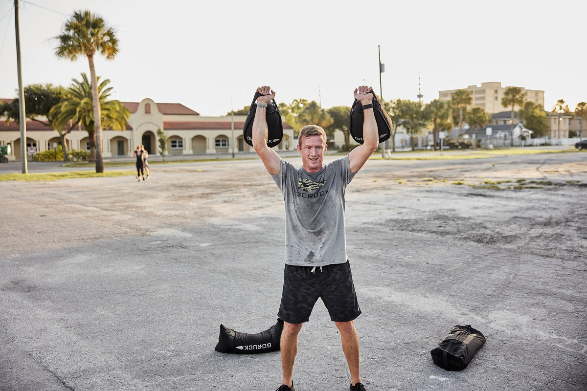 Man outdoors lifting GORUCK sandbags overhead, wearing GORUCK shirt, athletic training scene