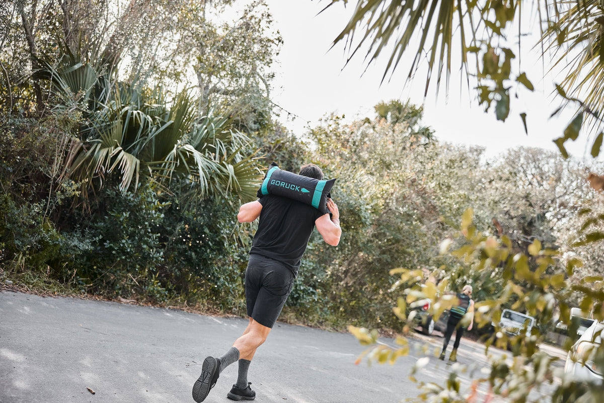 Man running outdoors carrying GORUCK sandbag, showcasing rucking fitness gear