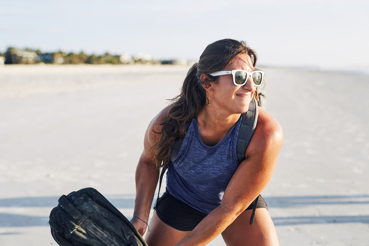 Woman in GORUCK gear exercising on a sandy beach, wearing sunglasses and a backpack