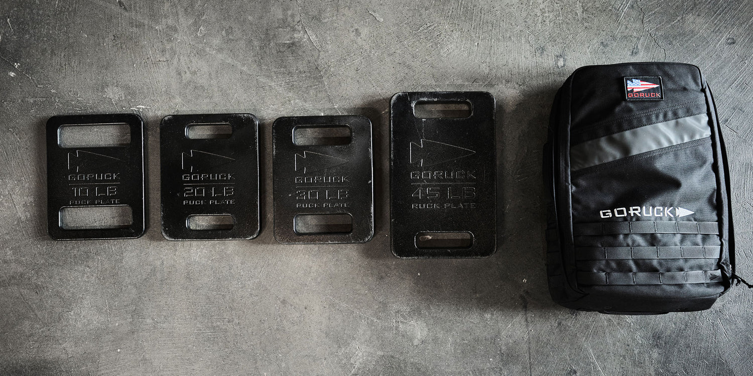 Four GORUCK ruck plates of varying weights next to a black GORUCK backpack on a concrete floor.