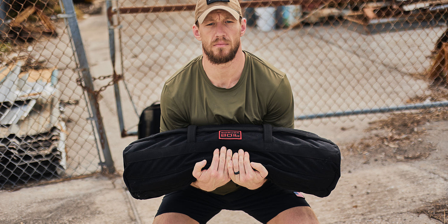 Man in a cap lifting a sandbag while squatting, next to a chain-link fence outdoors.