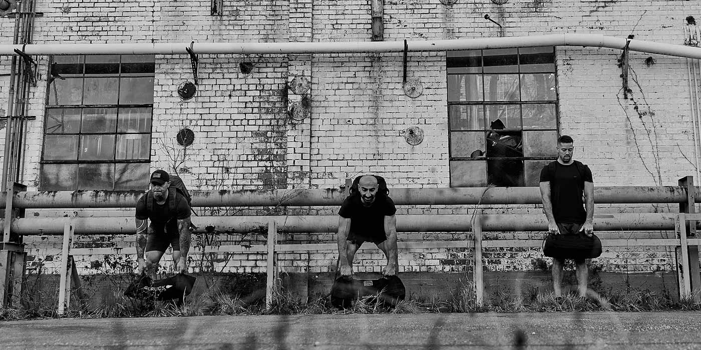 Three people exercise with sandbags in front of an old brick building with barred windows and pipes. Black and white photo.