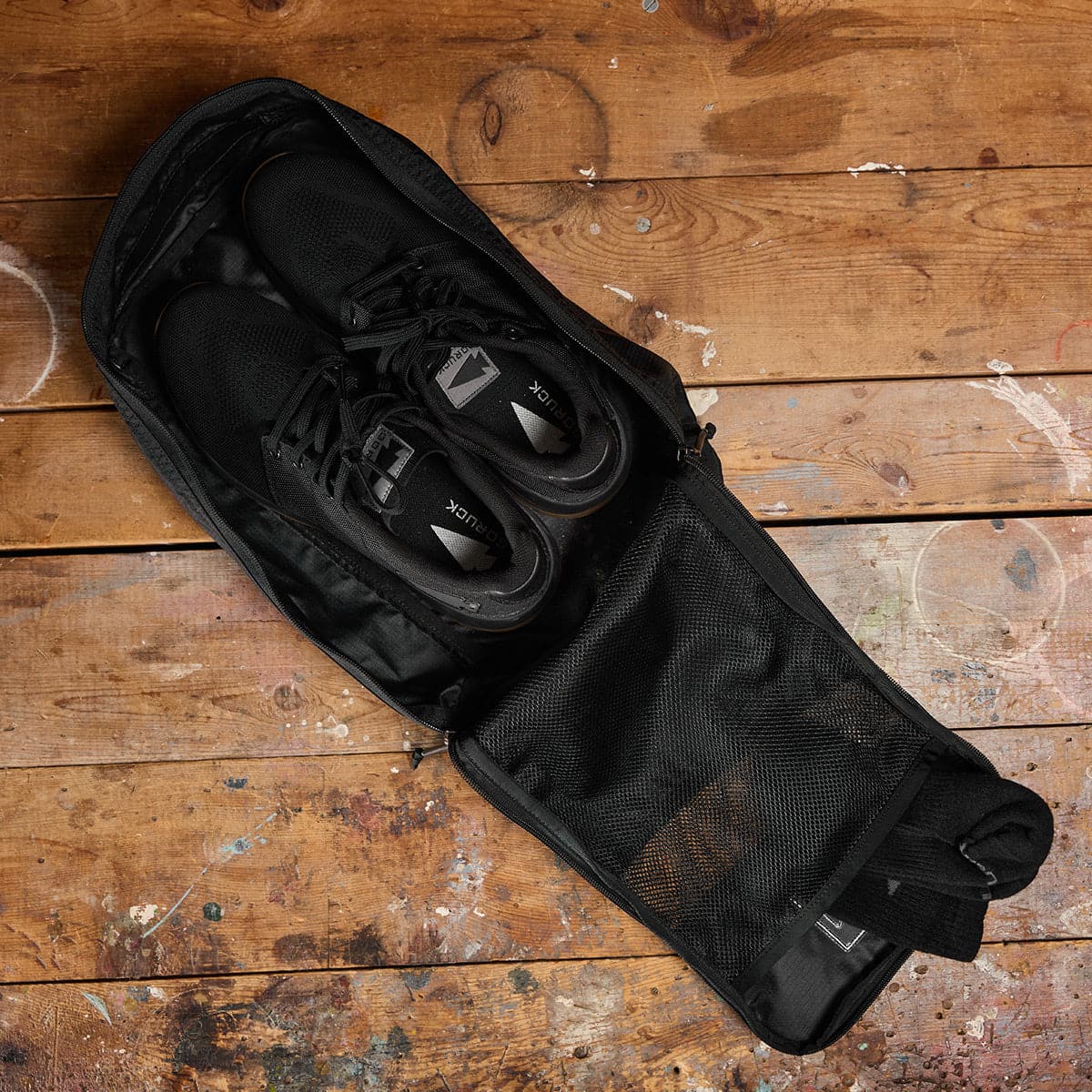 Shoes rest in a GORUCK Ripstop ROBIC shoe bag on a rustic wooden floor with paint splatters.