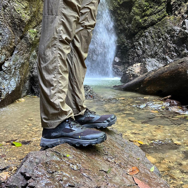 Hiker in tan pants and black boots stands on a rock by a stream and waterfall in a forest.