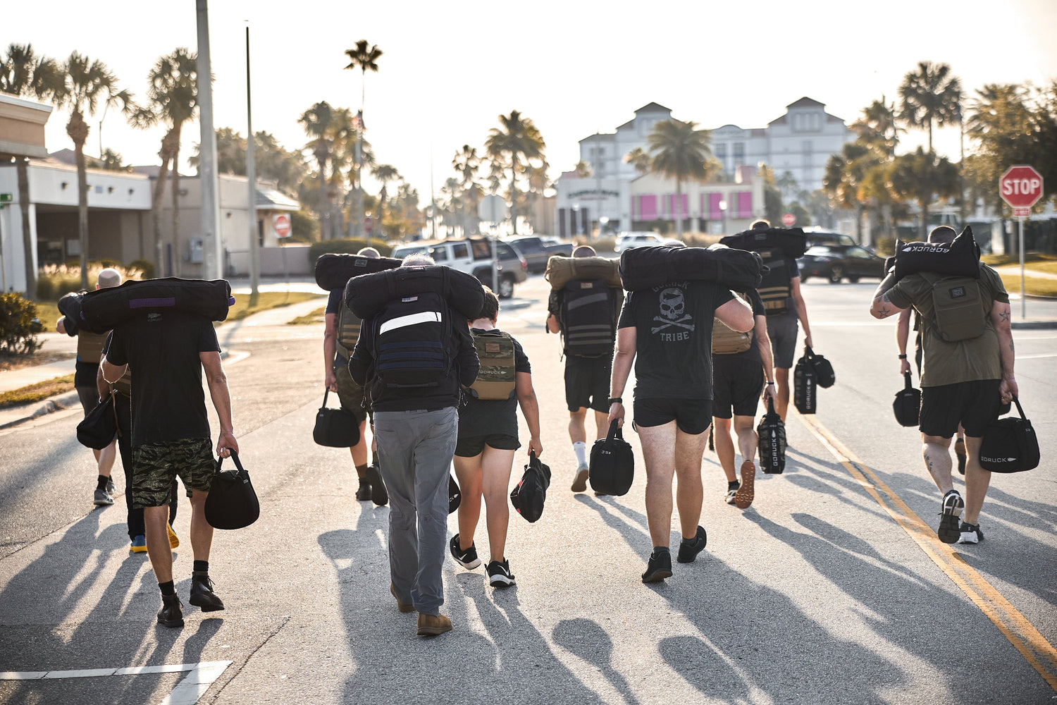 A group of people walk on a street carrying heavy backpacks and gear in bright, sunny weather.