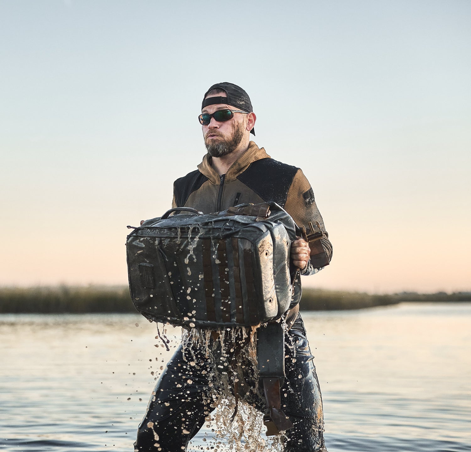 A man in sunglasses lifts a wet black bag from the water, with droplets splashing in the air.