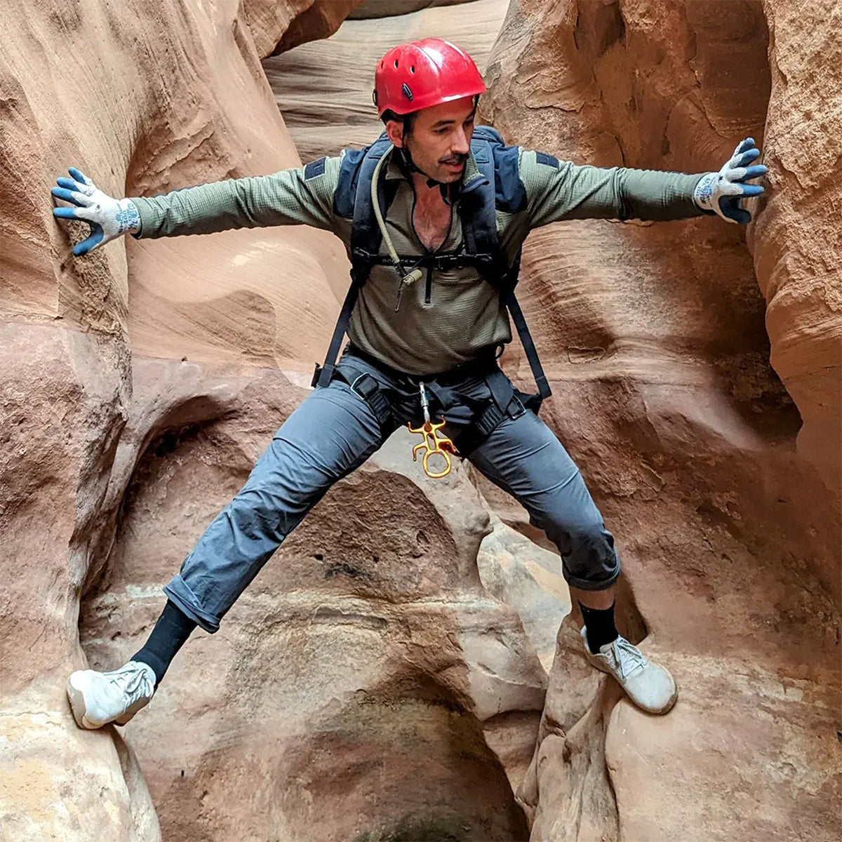 A man in climbing gear stretches across rock walls in a narrow canyon, using his hands and feet for support.