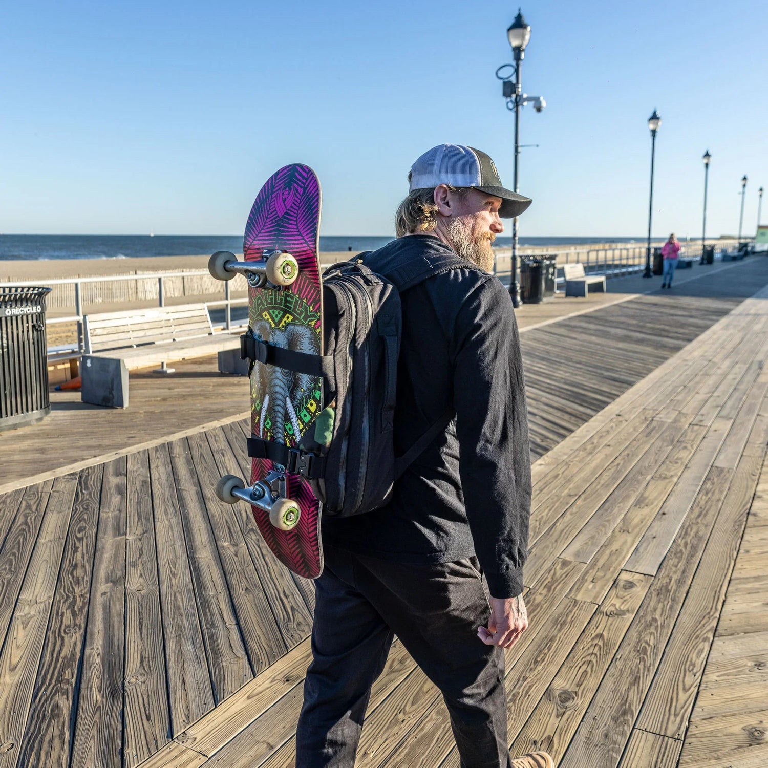 Man on boardwalk with skateboard attached to GORUCK backpack, ocean and benches in background