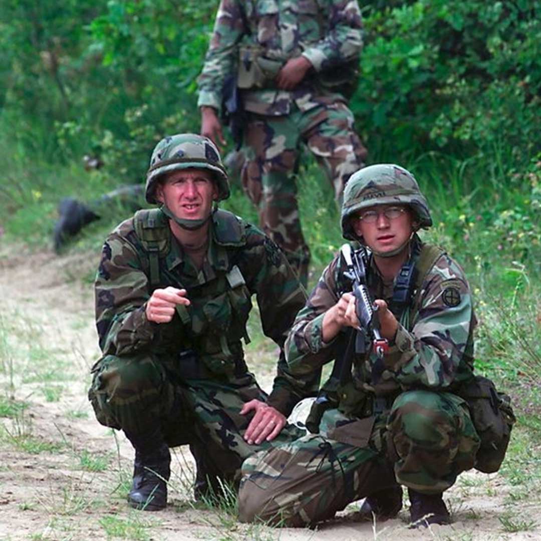 Two soldiers in camouflage kneel on a dirt path, one pointing, both wearing helmets and holding equipment.