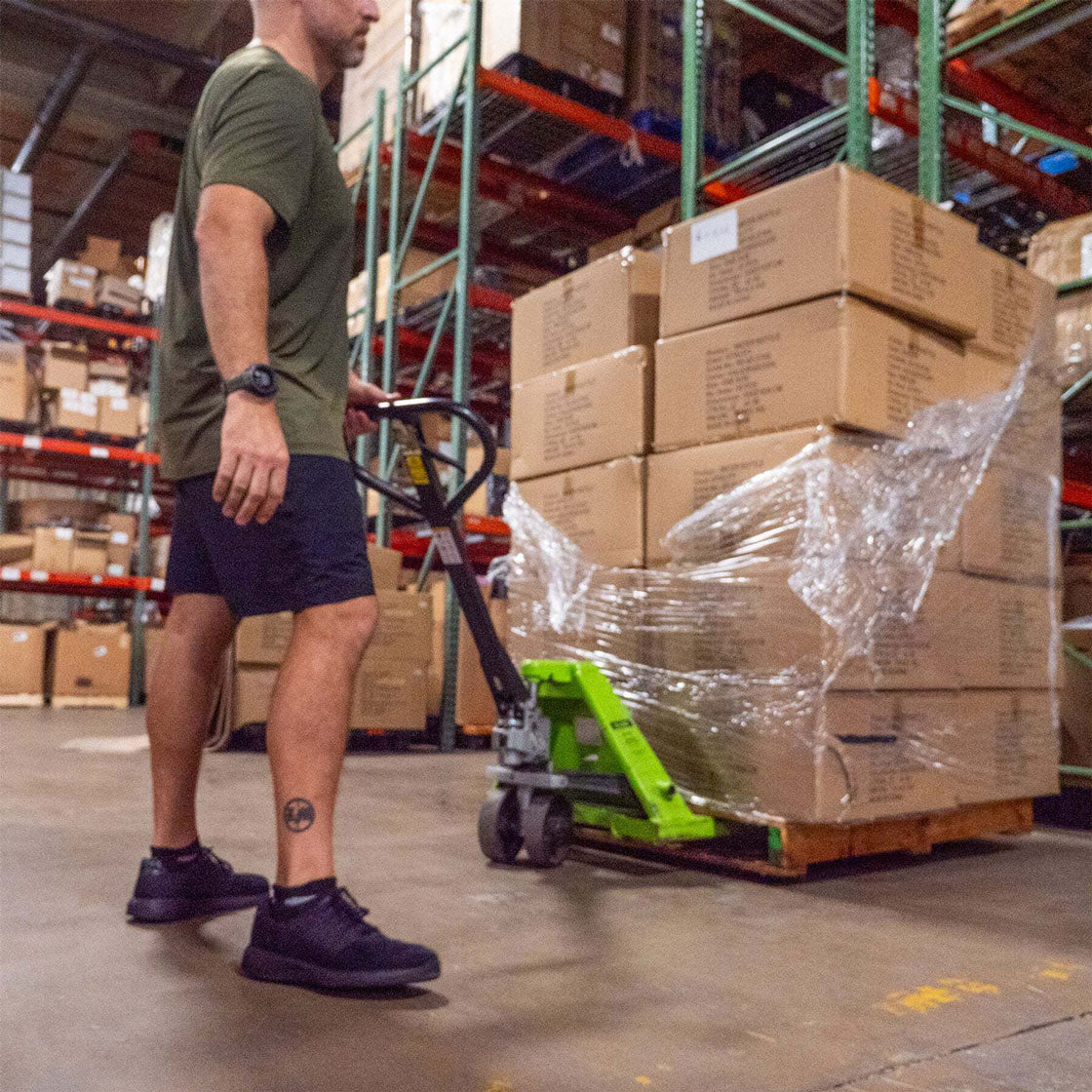 Person moving a pallet of boxed goods with a manual pallet jack in a warehouse.