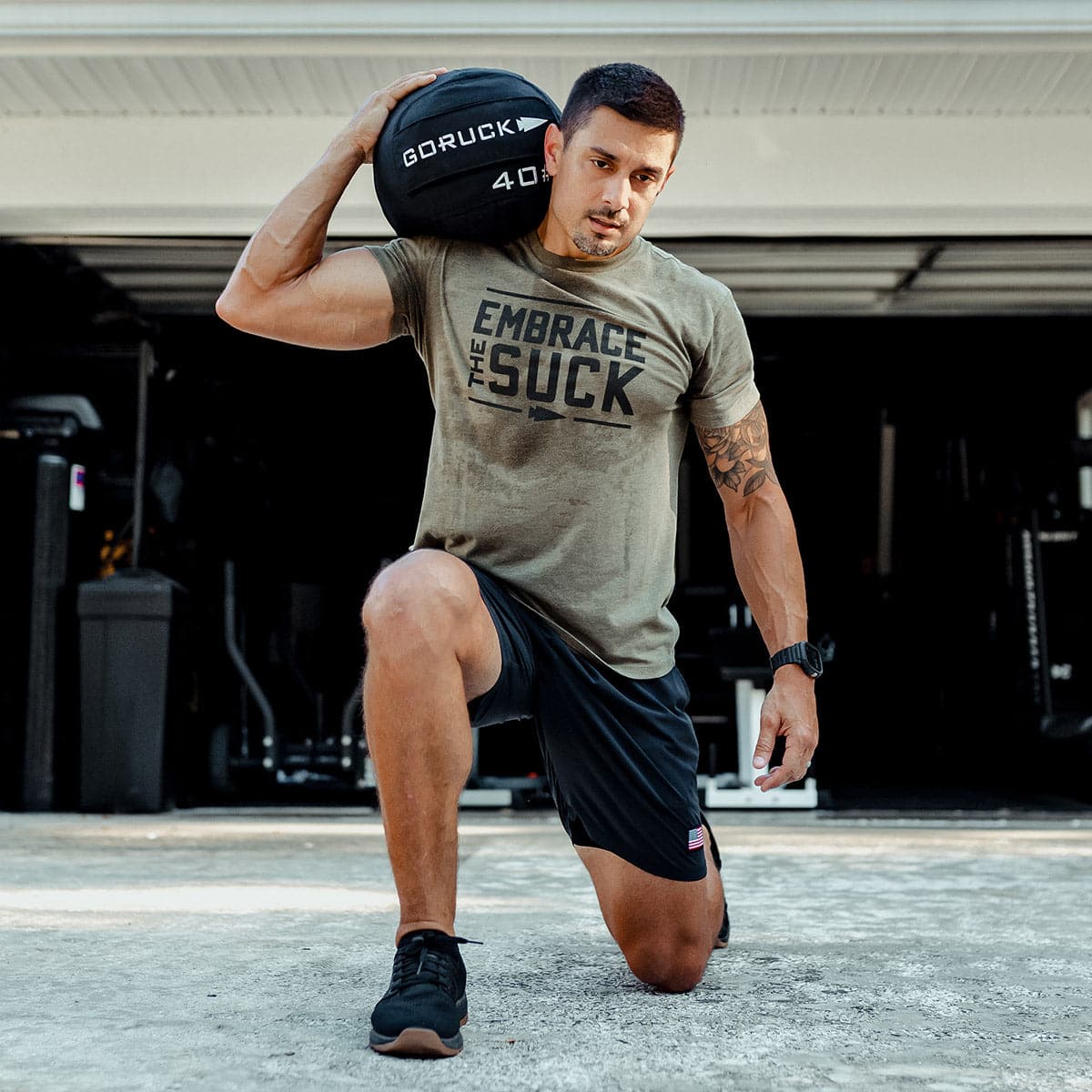 A man kneels on one leg wearing workout attire, holding a 40 lb Sand Medicine Ball by GORUCK on his shoulder and sporting an
