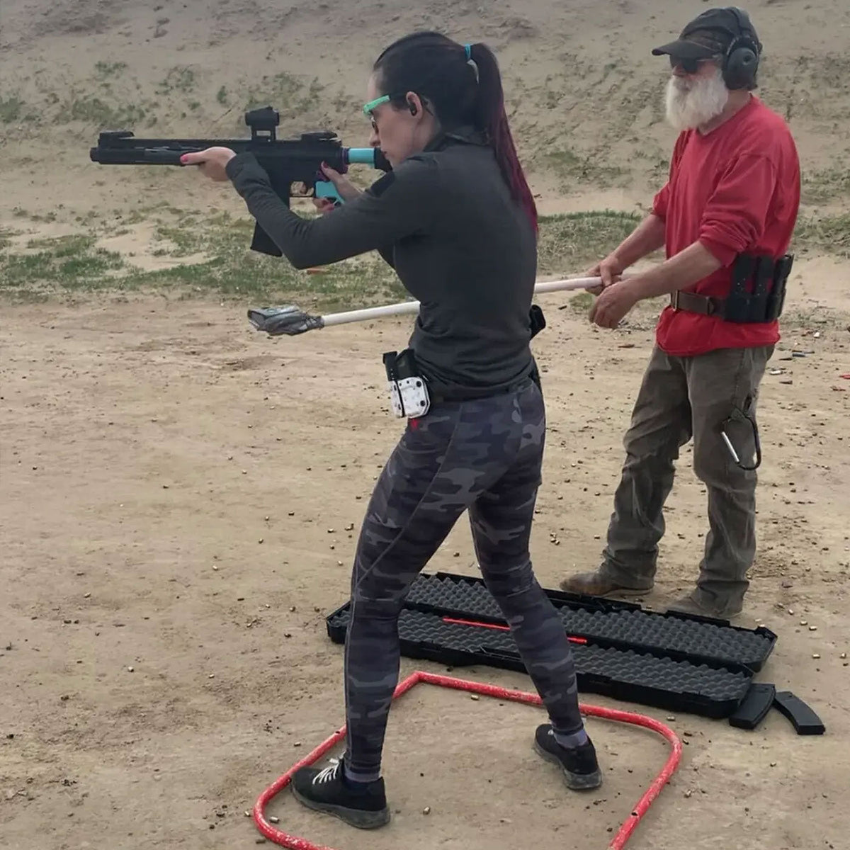Woman aiming a rifle at a shooting range, while a man in red shirt observes from behind.
