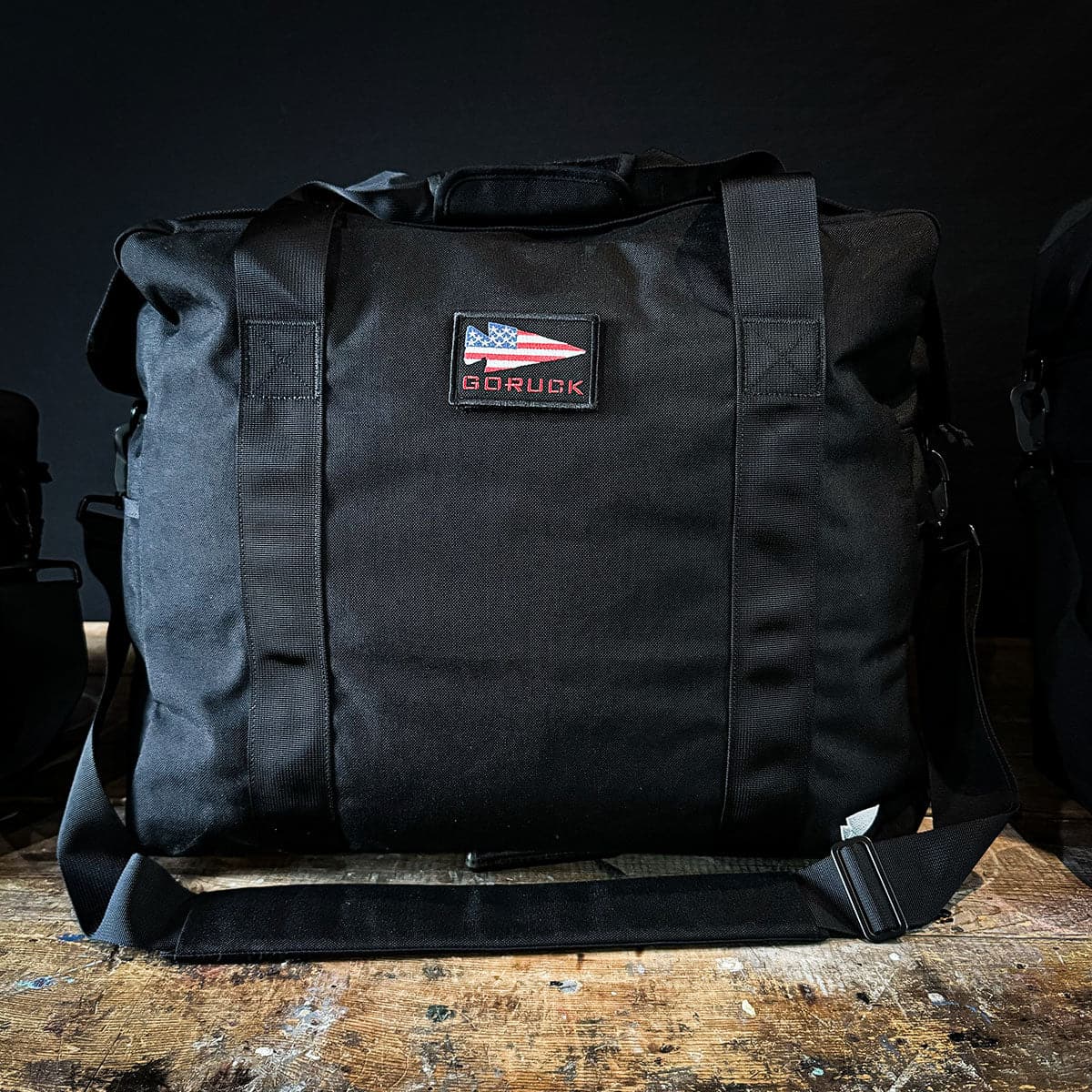 A black Kit Bag - 84L from GORUCK, featuring a design inspired by the Special Forces Flight Bag, is adorned with an American flag patch and rests on a wooden surface against a dark background.