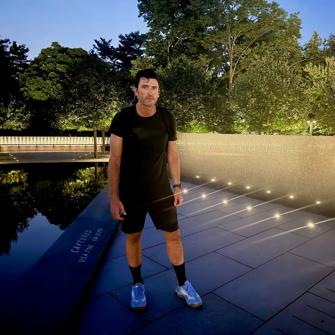 Wearing the Mackall 45 Americana, a man in a black outfit stands beside an illuminated memorial wall at night, where the words “FREEDOM IS NOT FREE” can be seen.