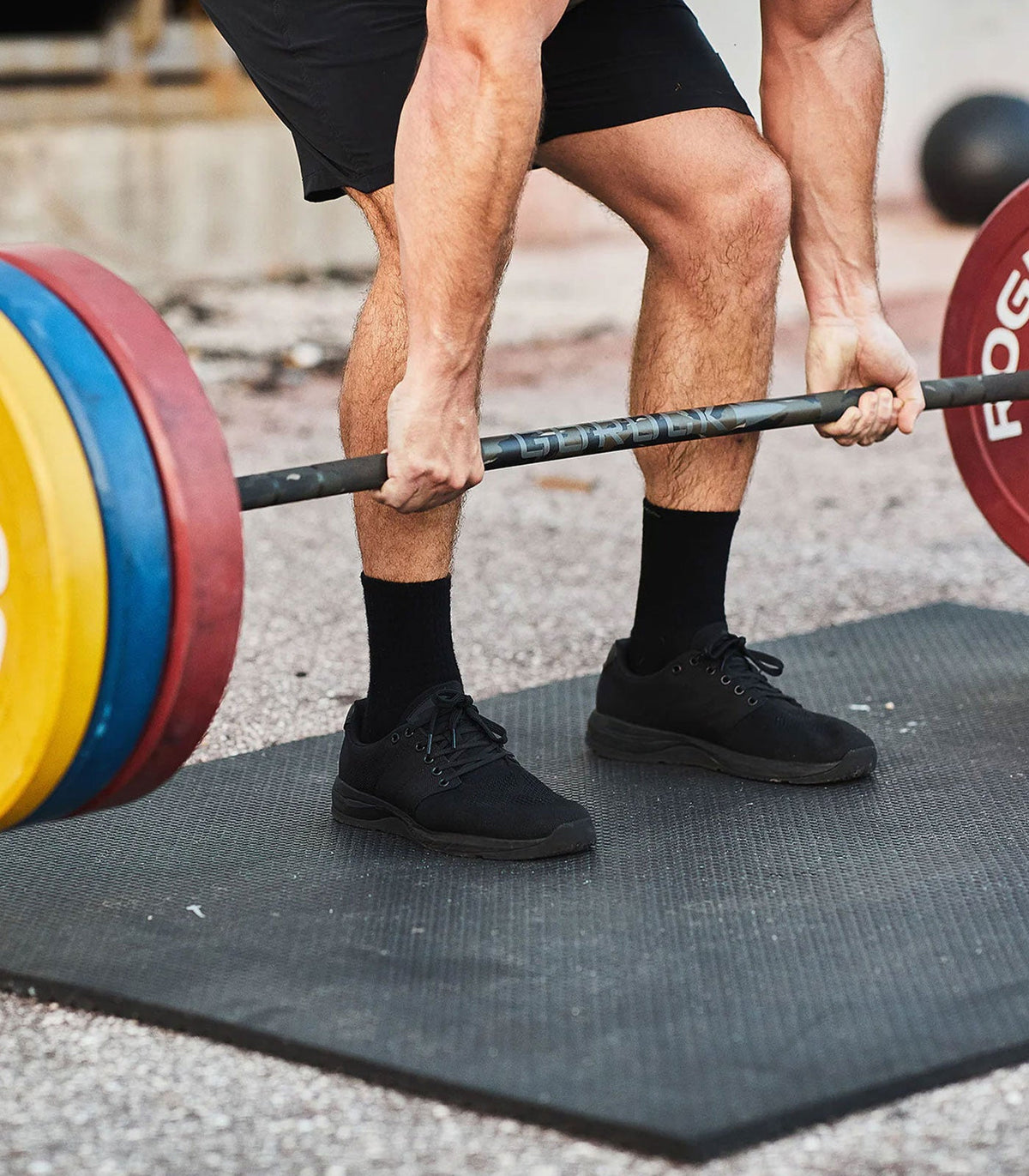 Person lifting a barbell with yellow, blue, and red weight plates on a black mat outdoors.