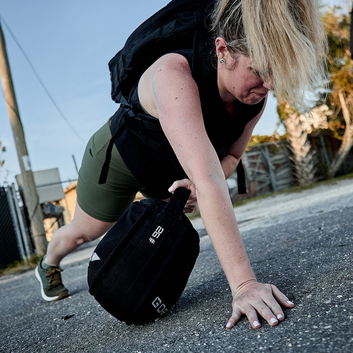 Woman doing an outdoor push-up, one hand on a weighted sandbag, wearing athletic gear and backpack.