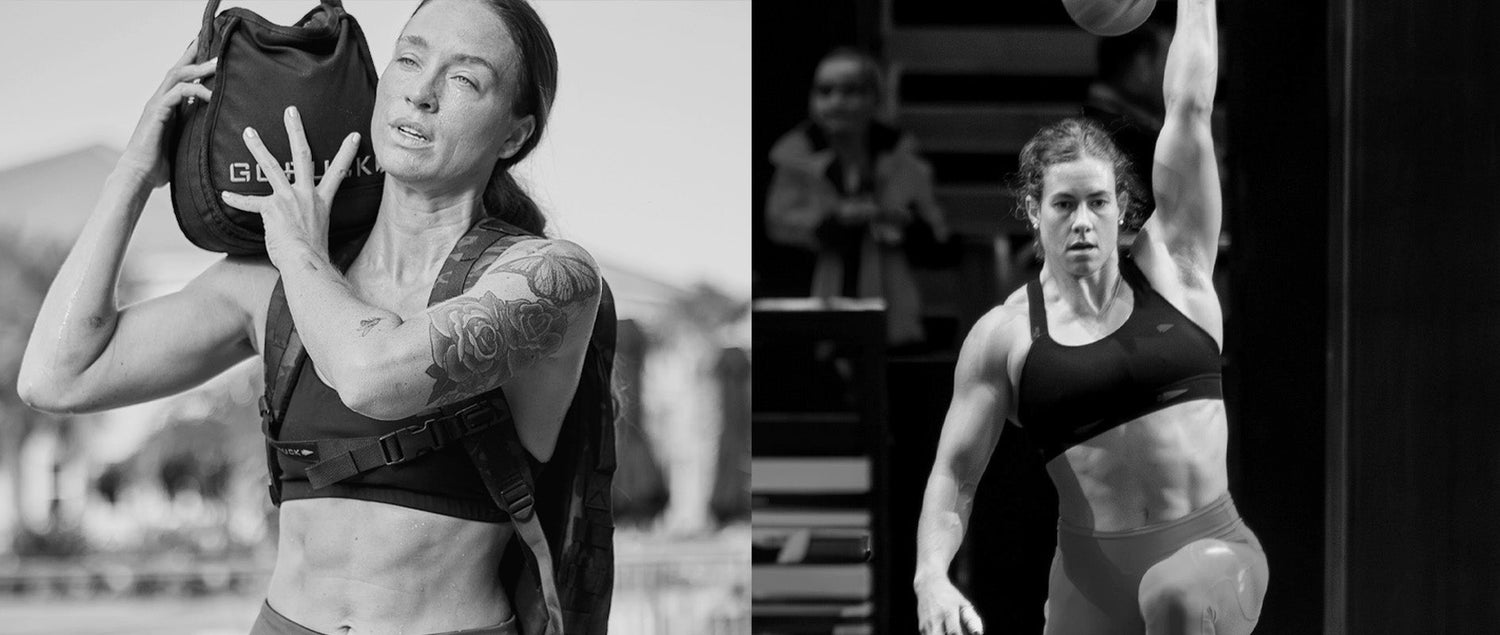 Two athletic women in sports bras training; one lifts a sandbag, the other lifts a kettlebell overhead.