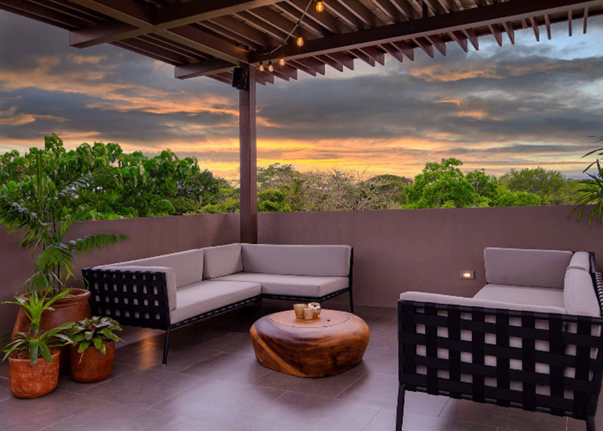 Modern patio with gray sofas, a wooden table, potted plants, and a sunset view over lush green trees.