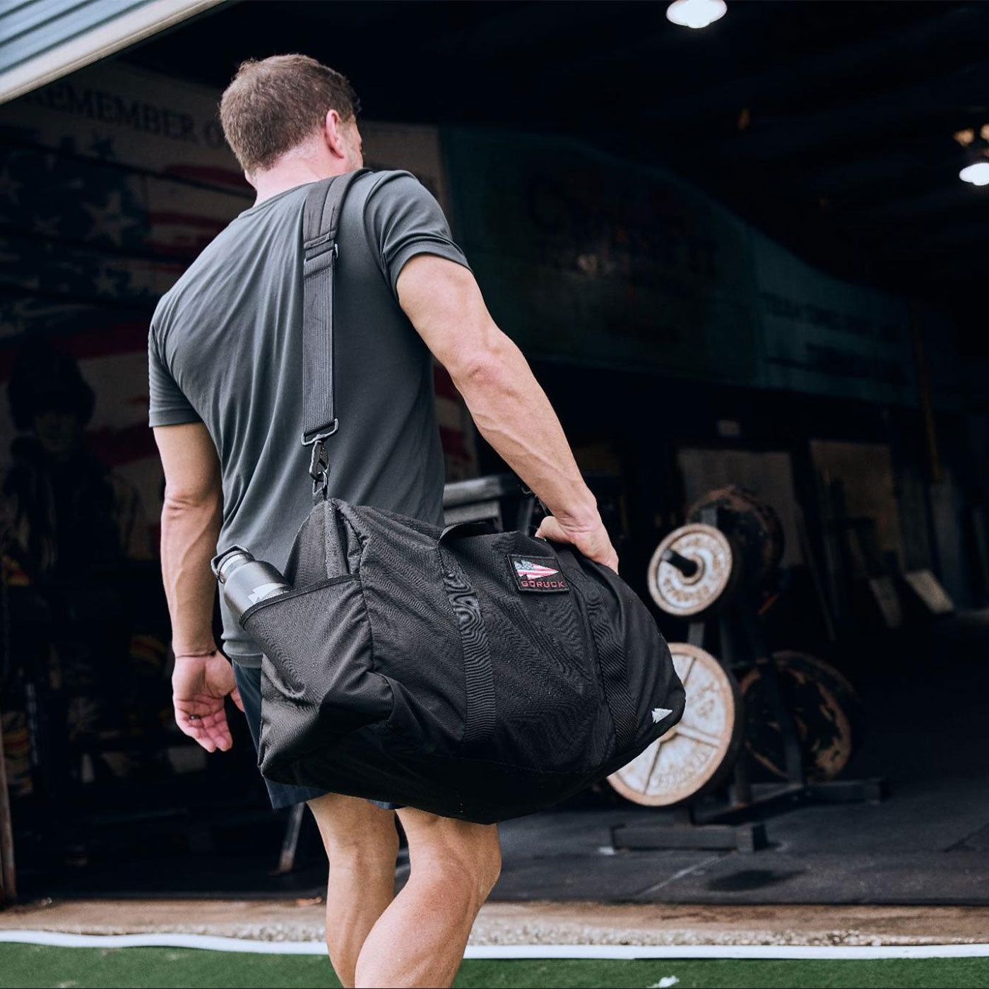 Man carrying a large black gym bag, walking toward a gym with weightlifting equipment inside.