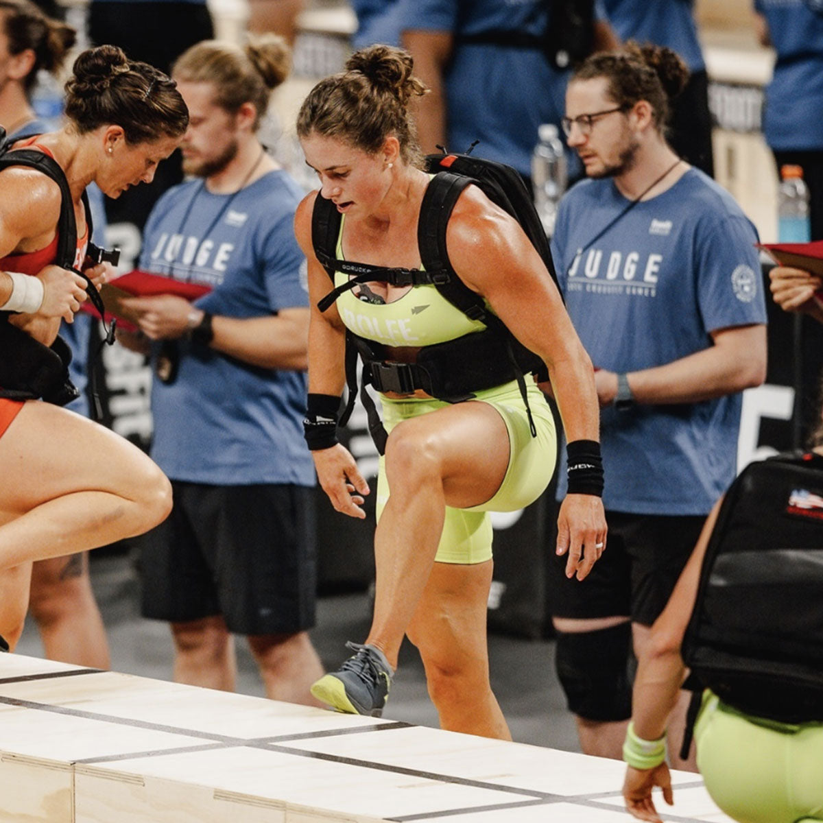 Athlete in yellow gear steps onto a wooden platform during a fitness competition, surrounded by judges.