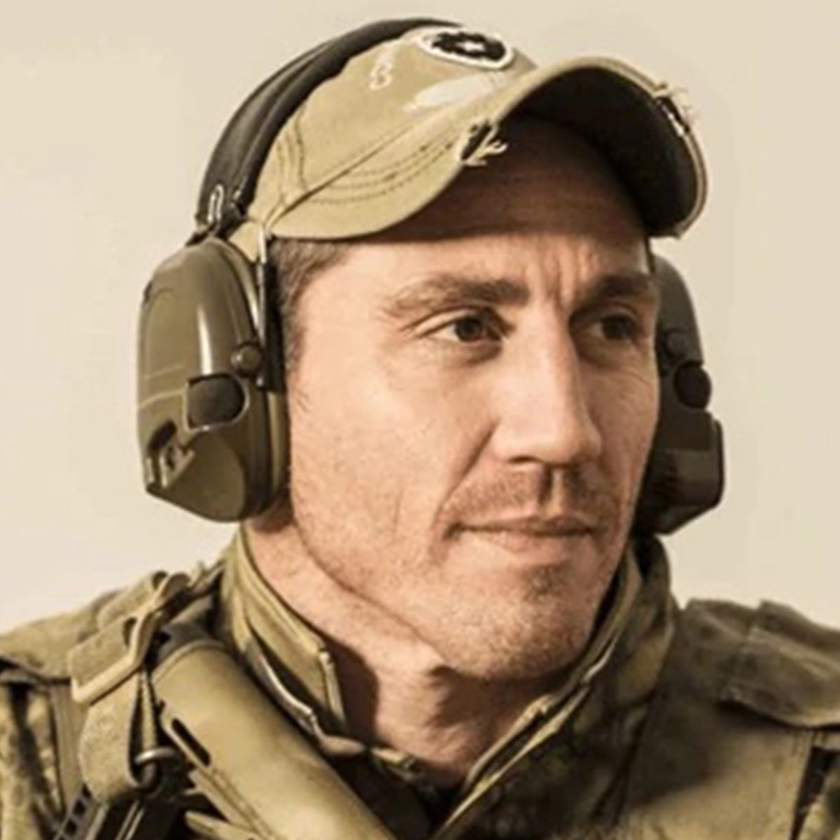 Man in military gear and a cap wearing tactical headphones, looking to the side with a neutral expression.