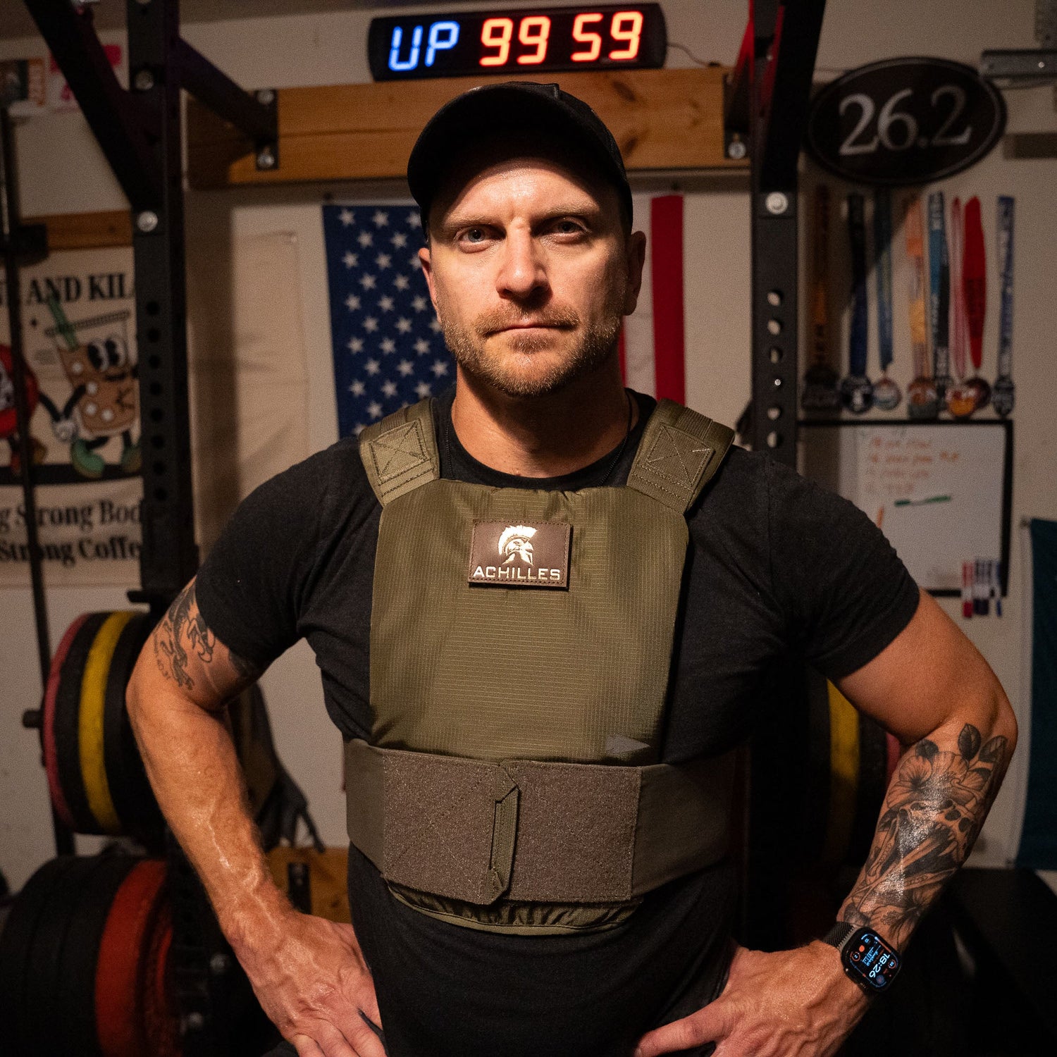 A man in a cap and Achilles Vest stands in a gym with an American flag and weights behind him.