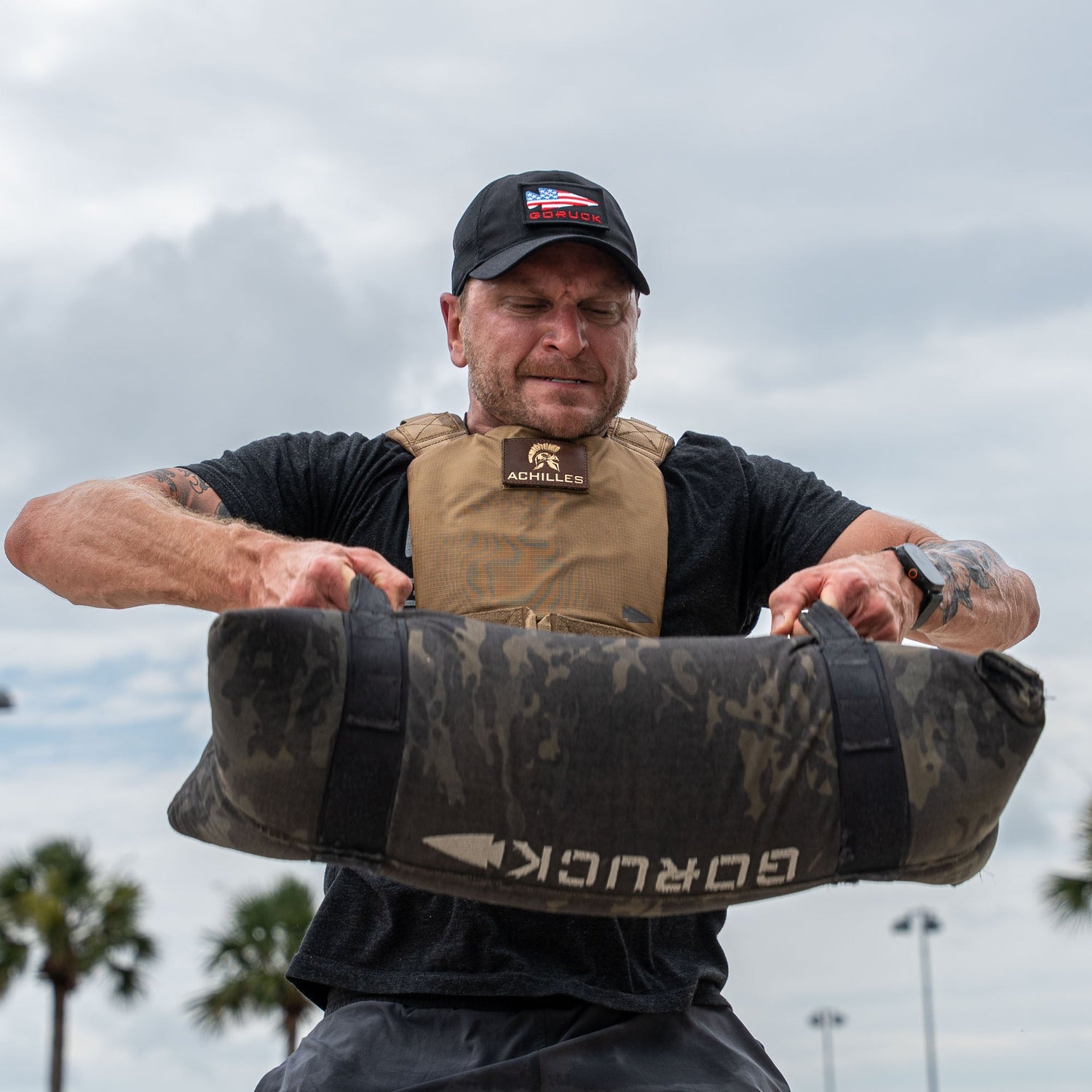 A man wearing the Achilles | Men's Weighted Vest lifts a heavy sandbag outdoors with palm trees and a cloudy sky in the background.