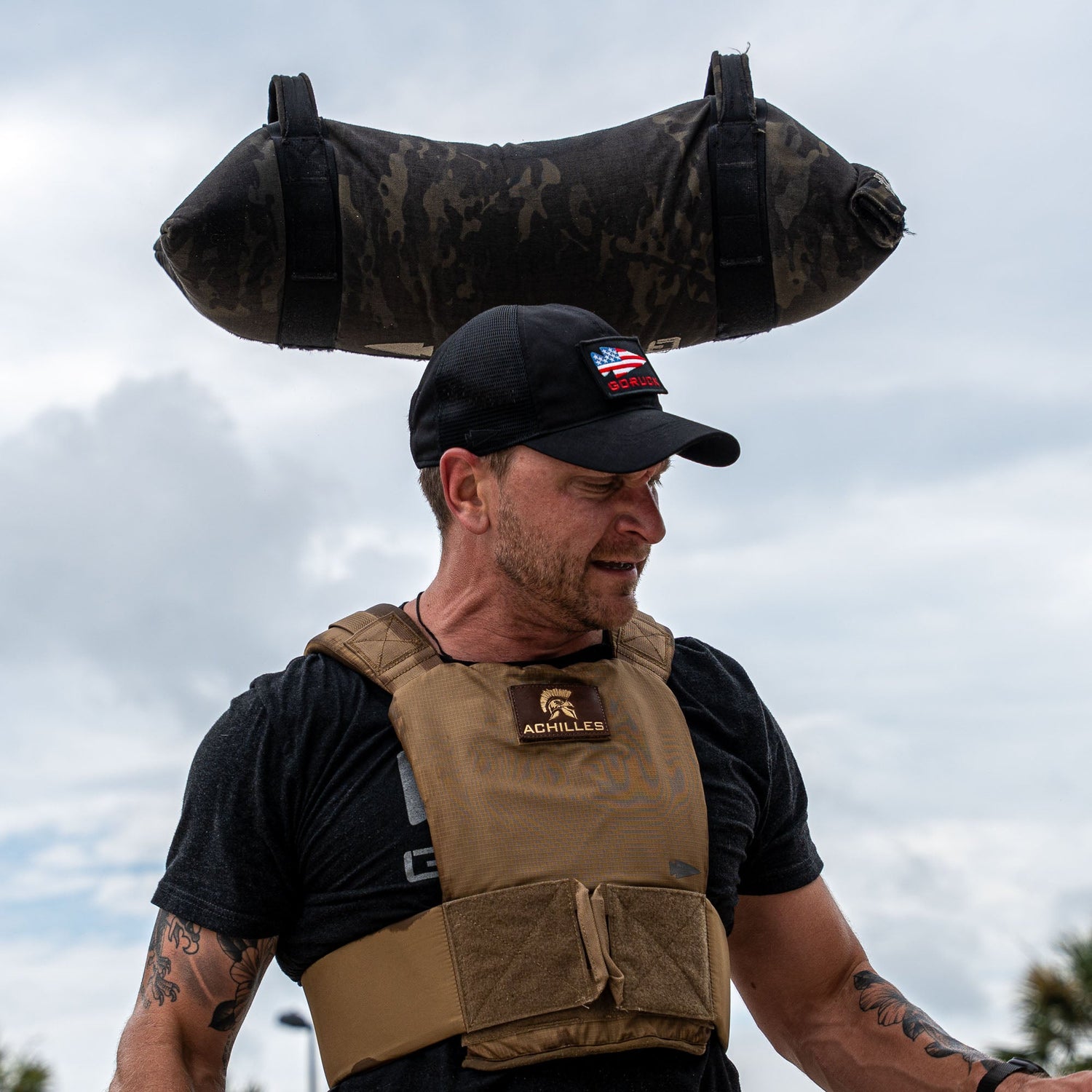 A man wears the Achilles | Men's Weighted Vest and a cap while carrying a large sandbag on his shoulders outdoors.