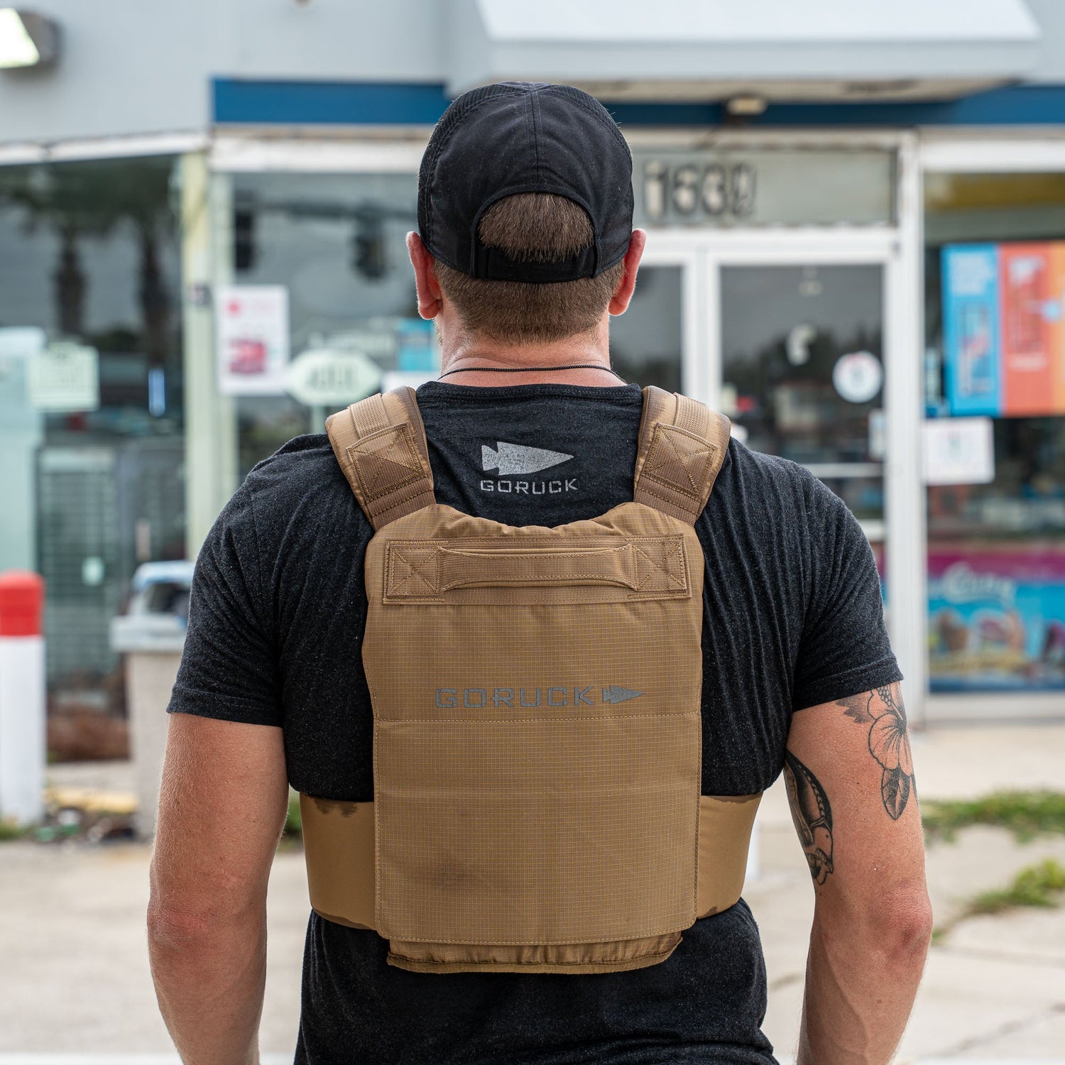 A man in a black cap and shirt wears the Achilles | Men's Weighted Vest in tan, standing outside a store with his back to the camera.