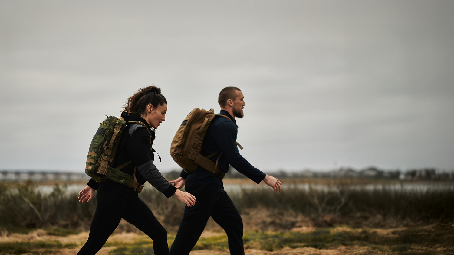 Two people wearing backpacks walk outdoors on a cloudy day near marshland and water.