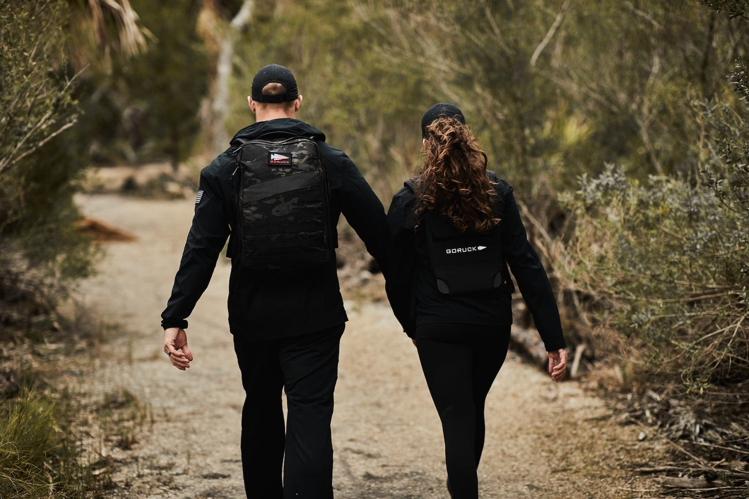 Two people in black outfits and backpacks walk hand-in-hand down a dirt trail surrounded by trees.