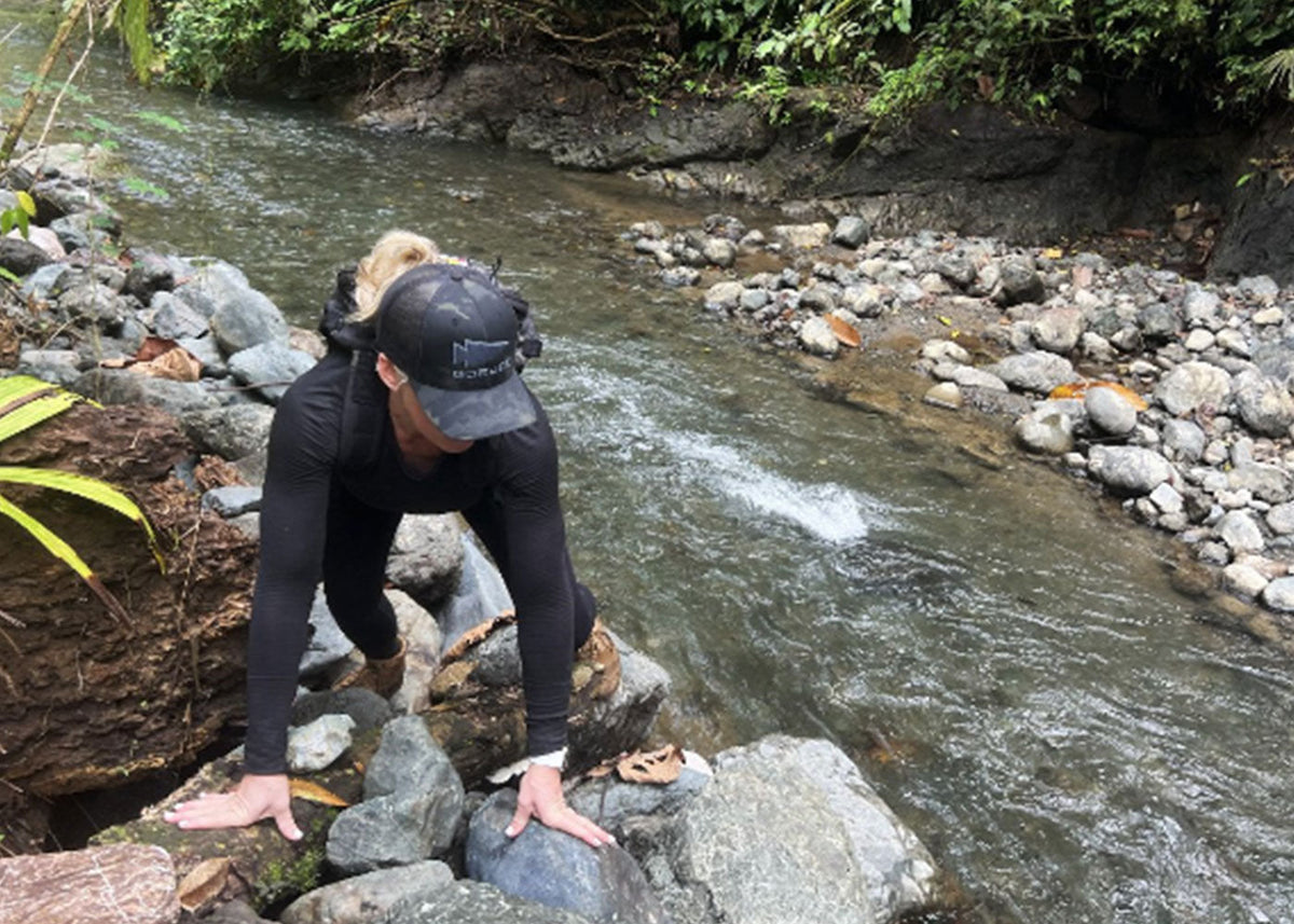 A person in black clothing climbs over rocks beside a flowing river in a lush, forested area.