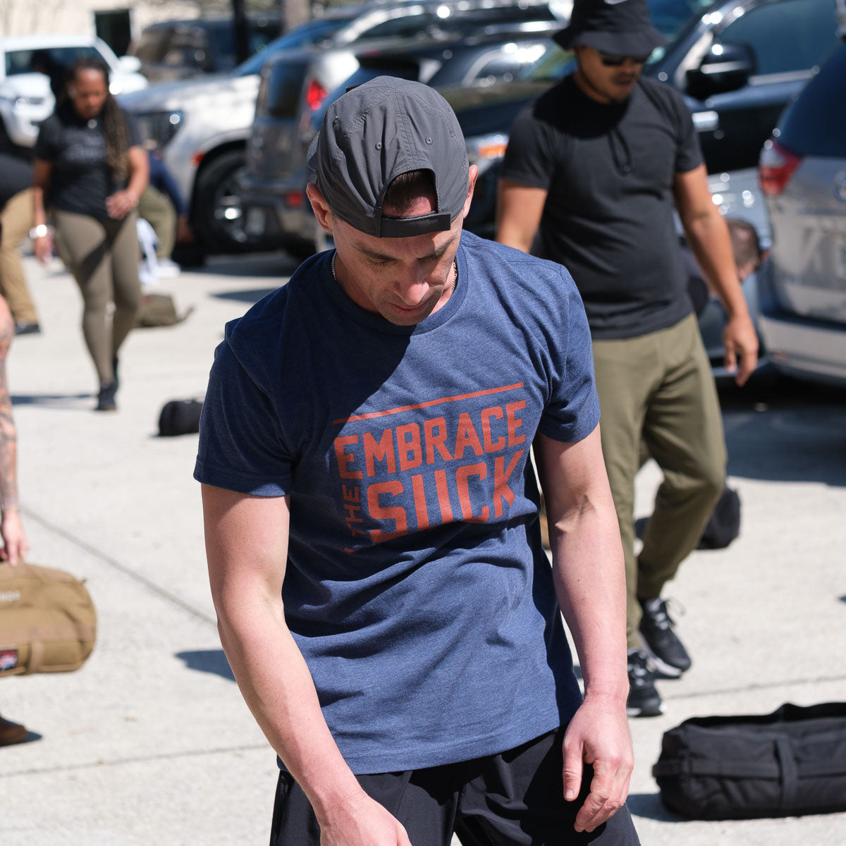 Man wearing navy GORUCK Embrace the Suck tee at outdoor rucking event with others.