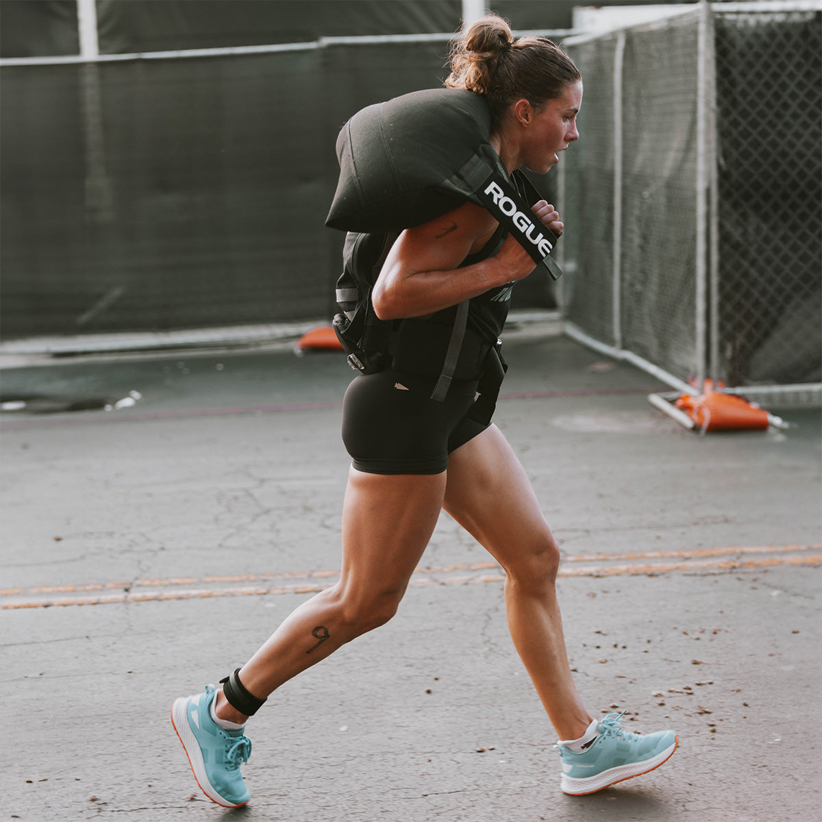 A woman runs outdoors carrying a large sandbag over her shoulders during a fitness event.