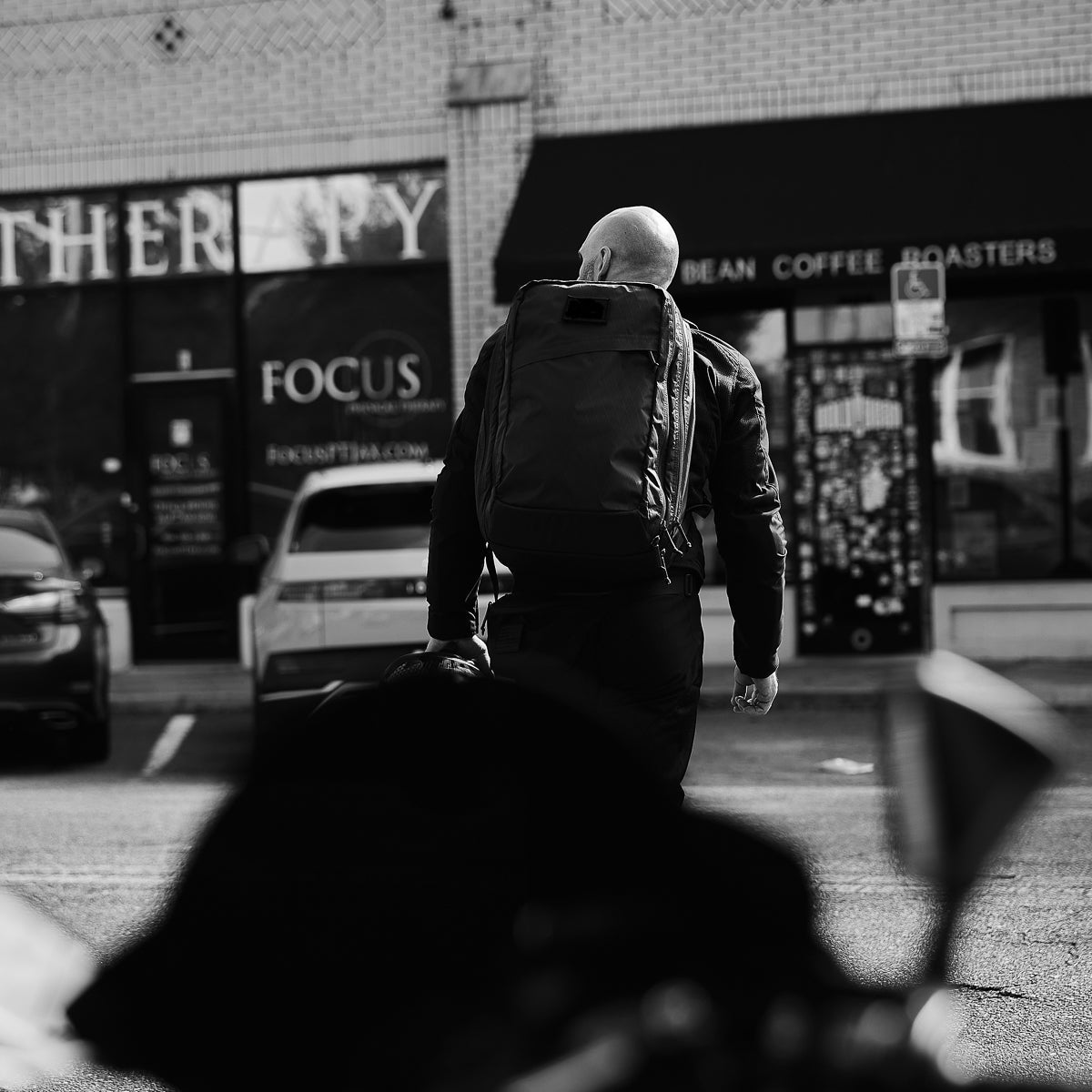 Man wearing GORUCK black backpack walking in urban street, parked cars and storefronts visible