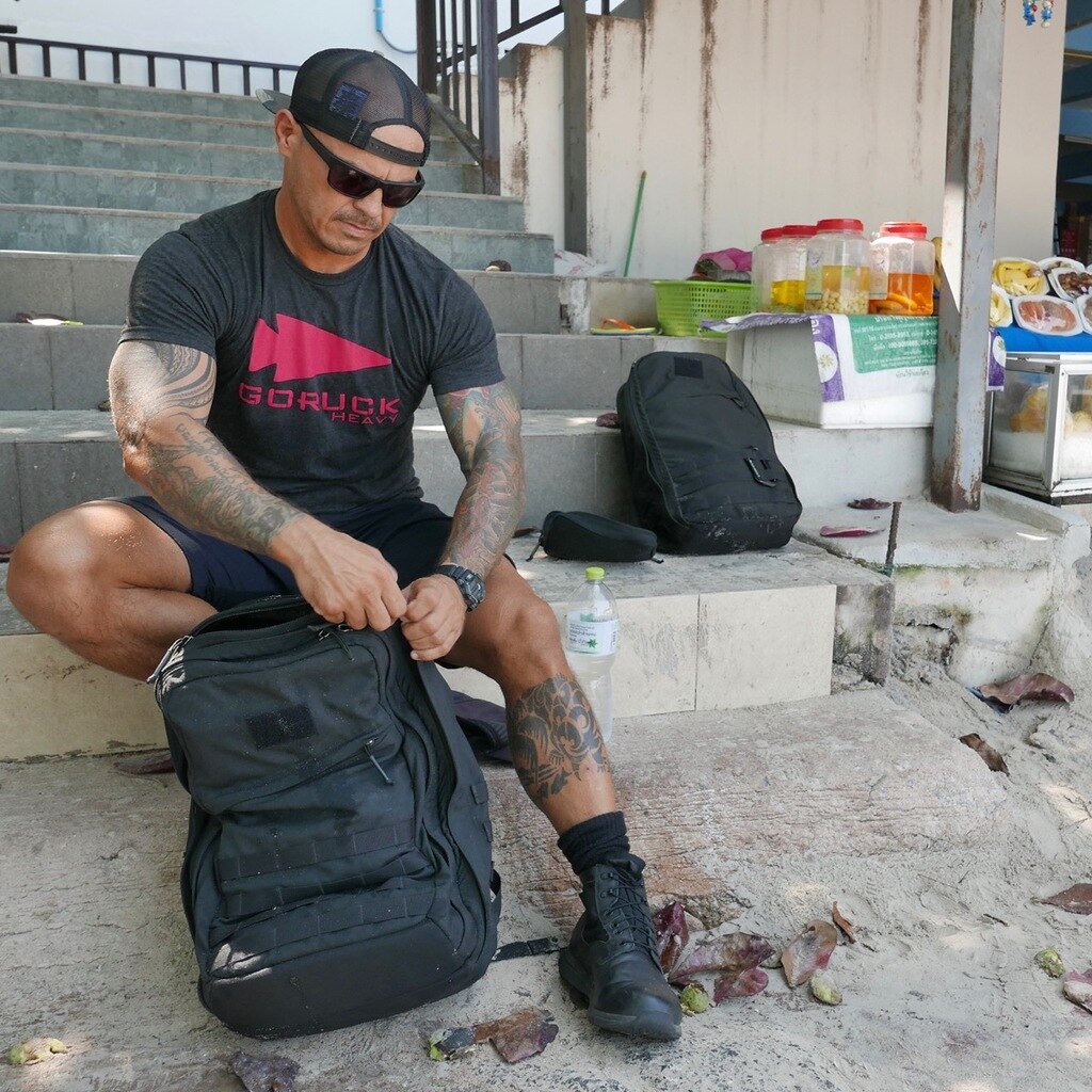 Man in a cap and sunglasses sits on steps, packing a black backpack, with snacks and drinks nearby.