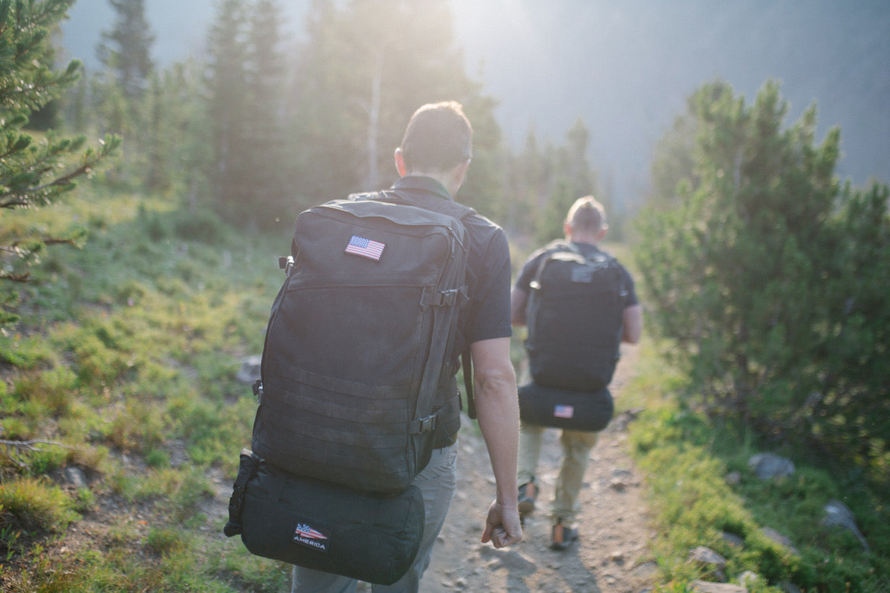 Two people with backpacks hike on a sunlit forest trail, heading away from the camera.